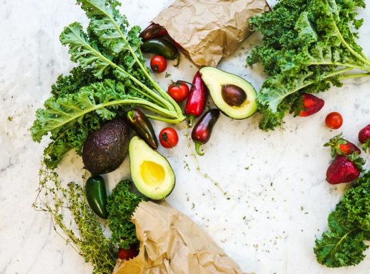 Avocado, kale, and more fresh produce artfully arranged on a marble surface.