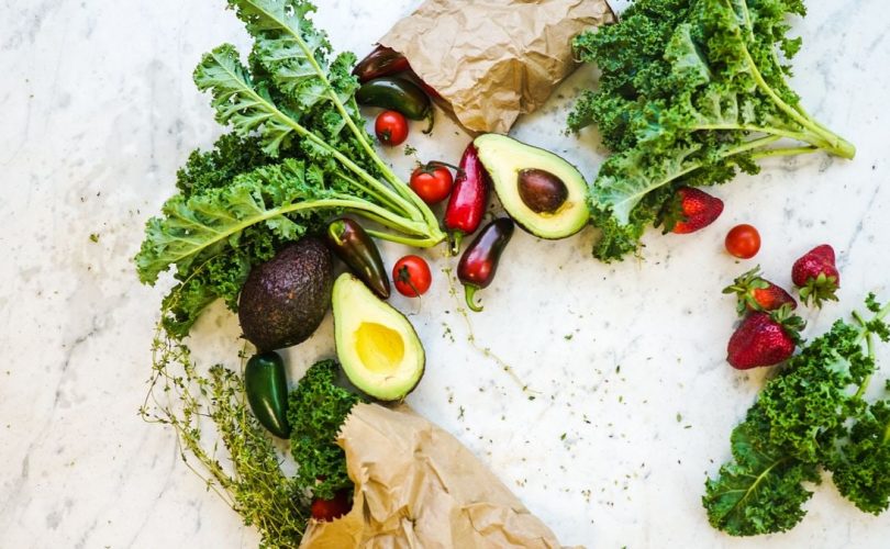 Avocado, kale, and more fresh produce artfully arranged on a marble surface.